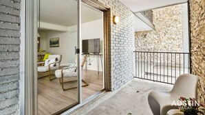 A modern apartment with a balcony, a stone wall, and a sliding glass door leading to a living area with a sofa and a TV. - Walk to Golf Course! Near Lake + Pool w Parking (Austin)