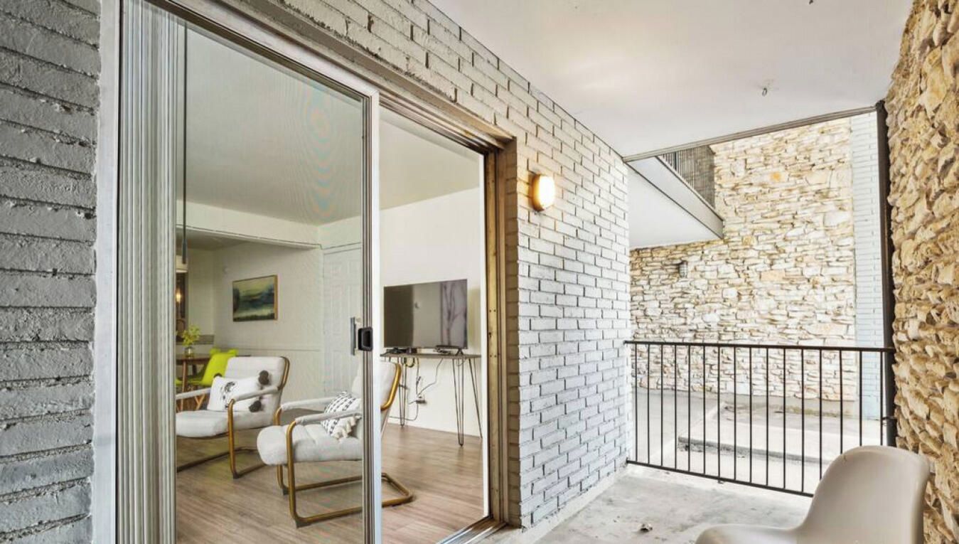 A modern interior with a glass door leading to a balcony, a seating area with a sofa and armchairs, and a stone wall.