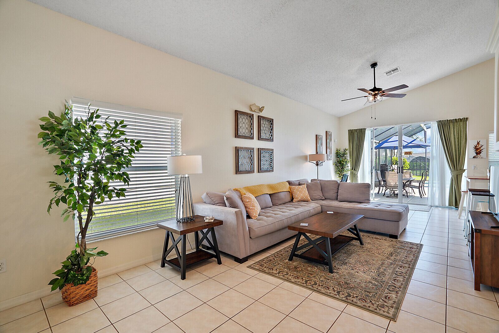 A spacious living room with a sectional sofa, a coffee table, a floor lamp, and a potted plant. There is a ceiling fan and a large window with blinds.