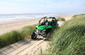 A green off-road vehicle on a sandy path beside a beach with ocean in the background.