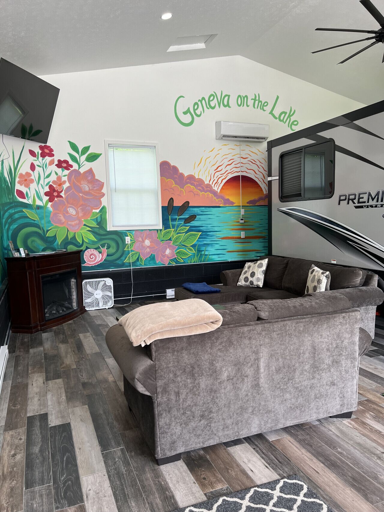 Un salon avec un canapé gris, une cheminée, une fresque murale intitulée « Genève au bord du lac » et un camping-car de modèle « FREMONT ULTRA ».