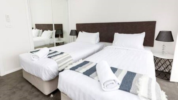 A hotel room with two beds, a large mirror, bedside lamps, and a neatly made bed with a striped blanket. - Escape to stylish comfort in this modern apartment! (Wagga Wagga)