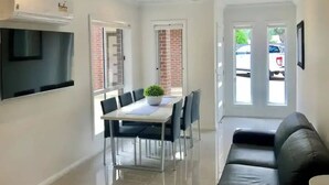 A modern living room with a black leather sofa, a dining area with a table and chairs, and a TV mounted on the wall.