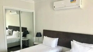 A hotel room with two beds, a mirror, a nightstand with a lamp, and an air conditioning unit.