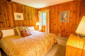 A wooden-paneled bedroom with a large bed, a nightstand, a lamp, and a framed picture on the wall. - Whiffletree D3 (Killington)