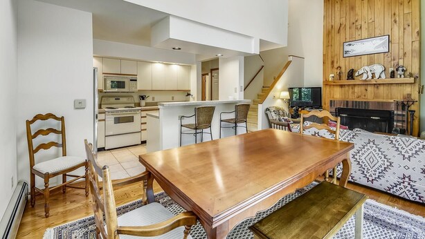 Ampla sala de estar com uma mesa de jantar de madeira, duas cadeiras, um sofá e uma televisão. Uma cozinha visível ao fundo, com eletrodomésticos modernos.