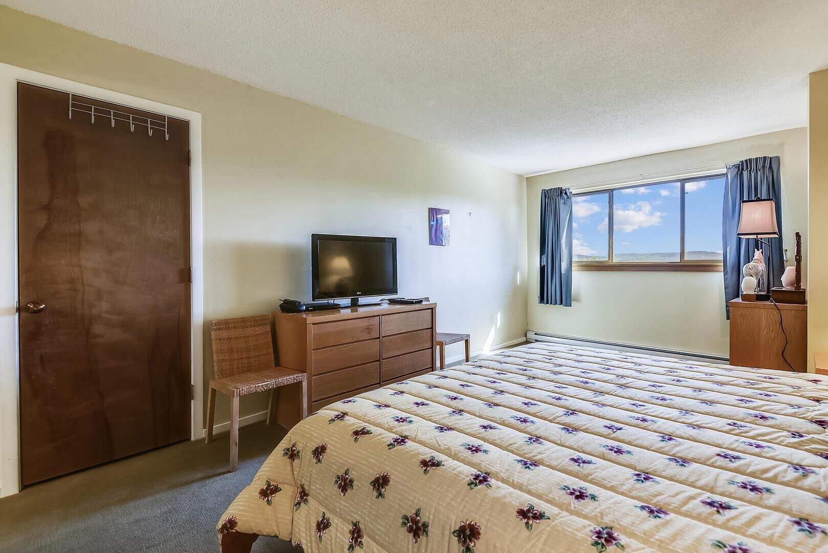 A bedroom with a bed, a television on a dresser, a chair, a window with curtains, and a door.