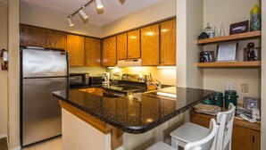 A compact kitchen with wooden cabinets, a stainless steel refrigerator, a microwave, and a sink. There are two white chairs and a small shelf with decorative items. - Pinnacle F21 (Killington)