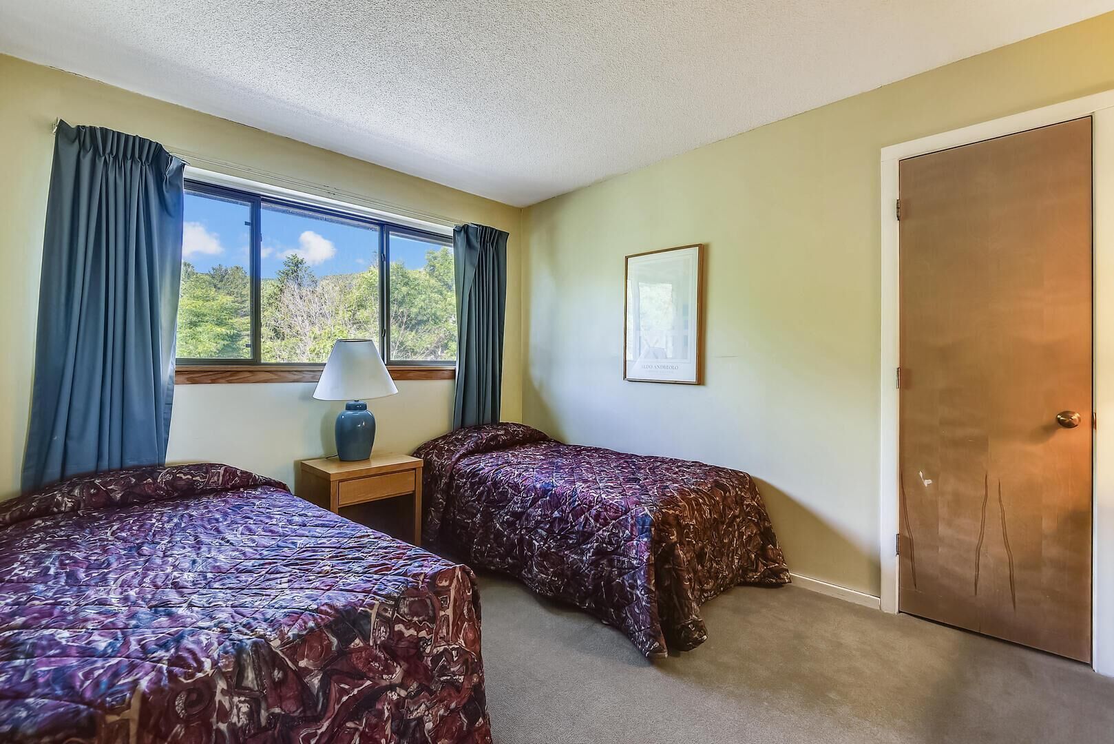 A room with two beds, a nightstand with a lamp, a framed picture on the wall, and a door.