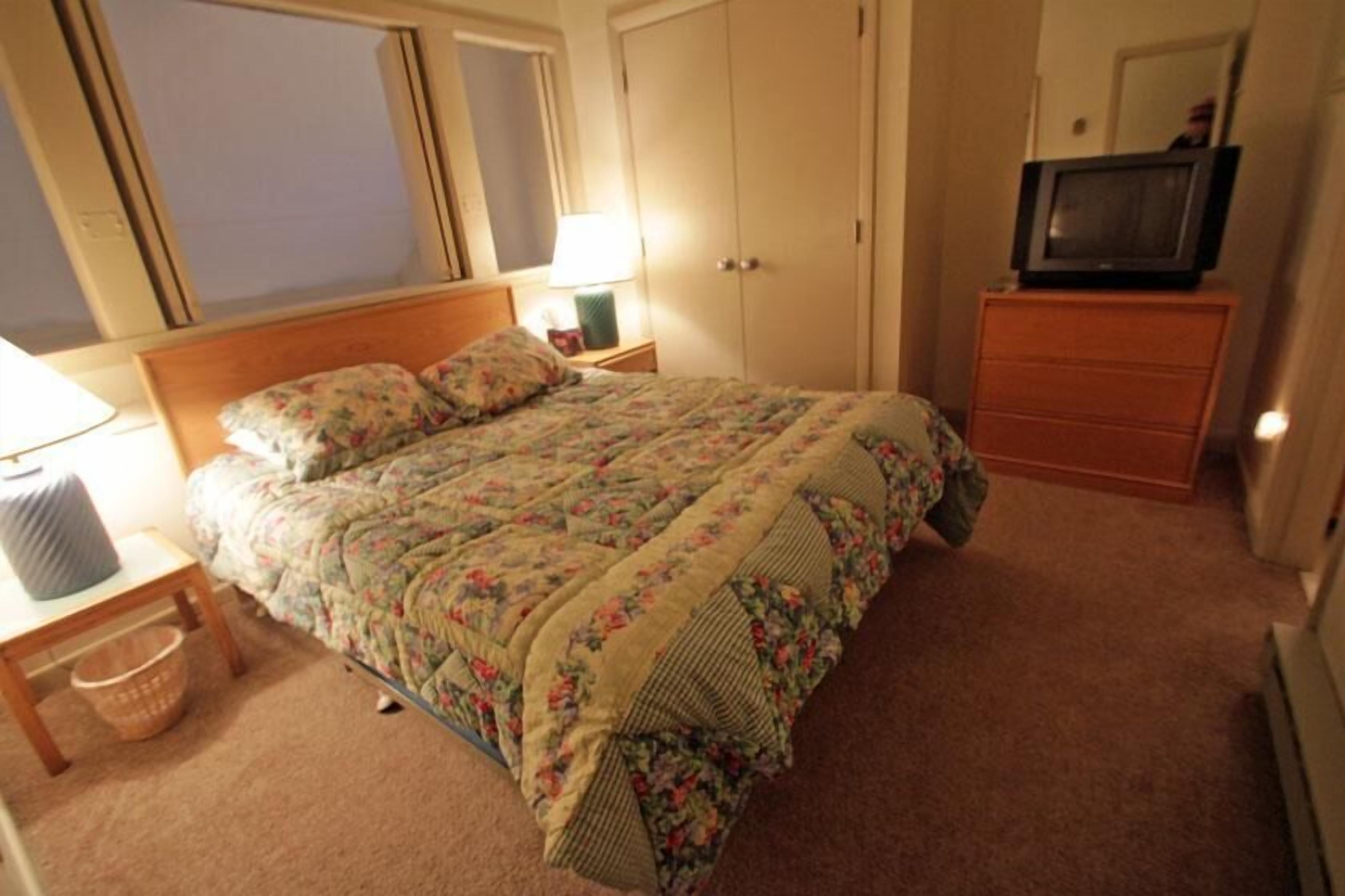 A bedroom with a bed, a dresser, a television on top of the dresser, and a window with blinds.