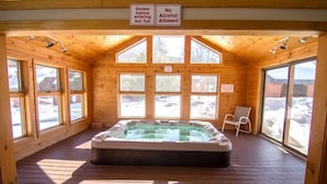 Una cabaña de madera con un jacuzzi, ventanas amplias y una silla. Señales indican ducharse antes de usar el jacuzzi y prohibición de alcohol.