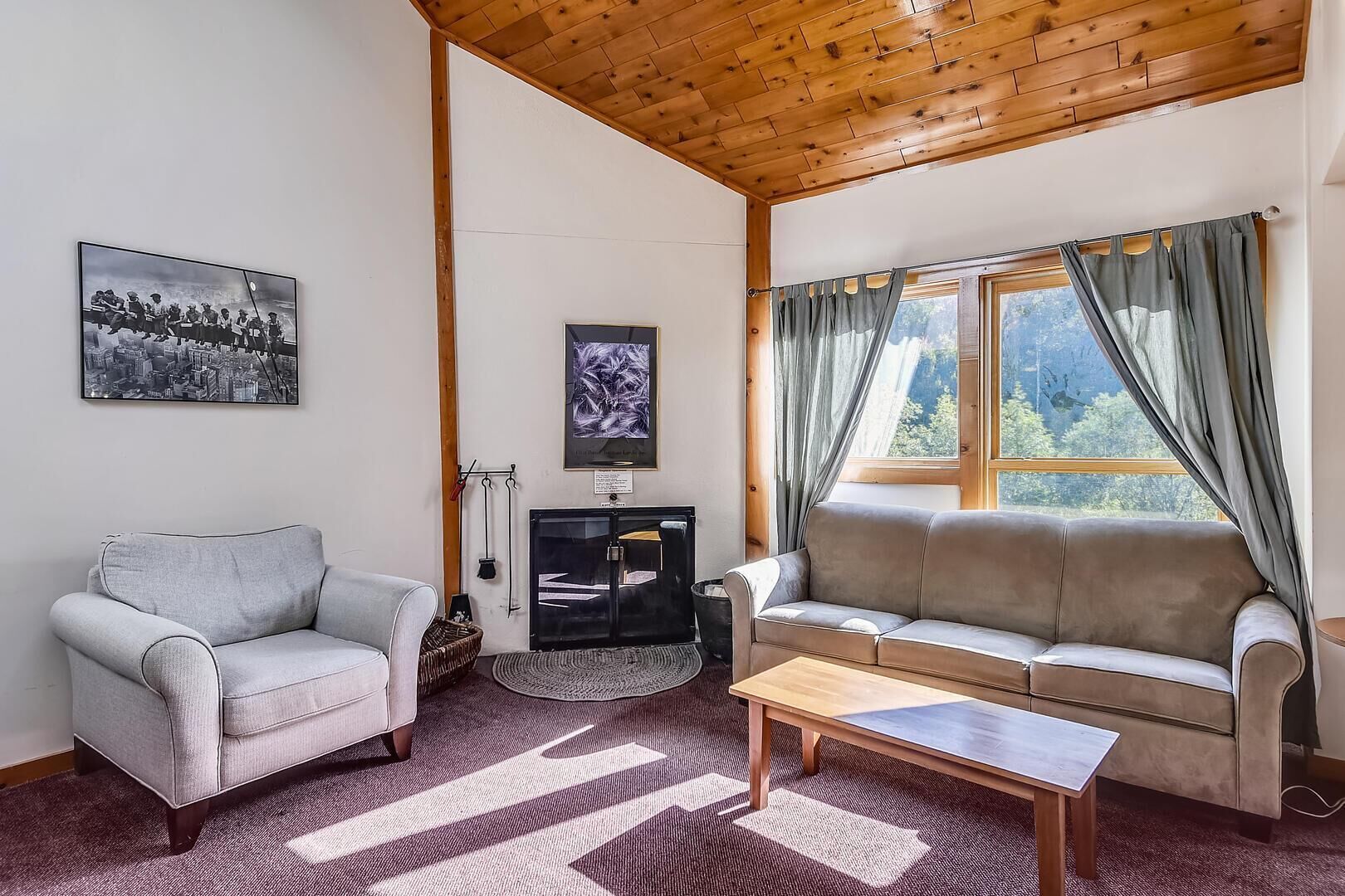 A cozy living room with a grey sofa, a grey armchair, and a wooden coffee table. There is a fireplace, a window with curtains, and artwork on the walls.