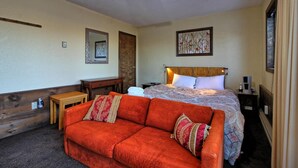 Habitación de hotel con una cama, cabecera de madera, sofá rojo, una mesita, un espejo y un cuadro en la pared.