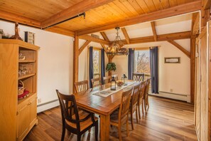 A dining area with a wooden table and chairs, a chandelier, and a view of trees through the windows. - Viewmont Chalet (Killington)
