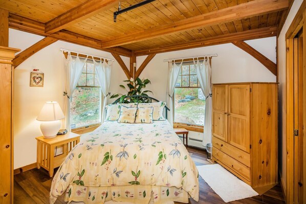 Une chambre avec un plafond en bois, un lit recouvert d’une literie à motifs floraux, une lampe de chevet et une vue sur l’extérieur par une fenêtre.