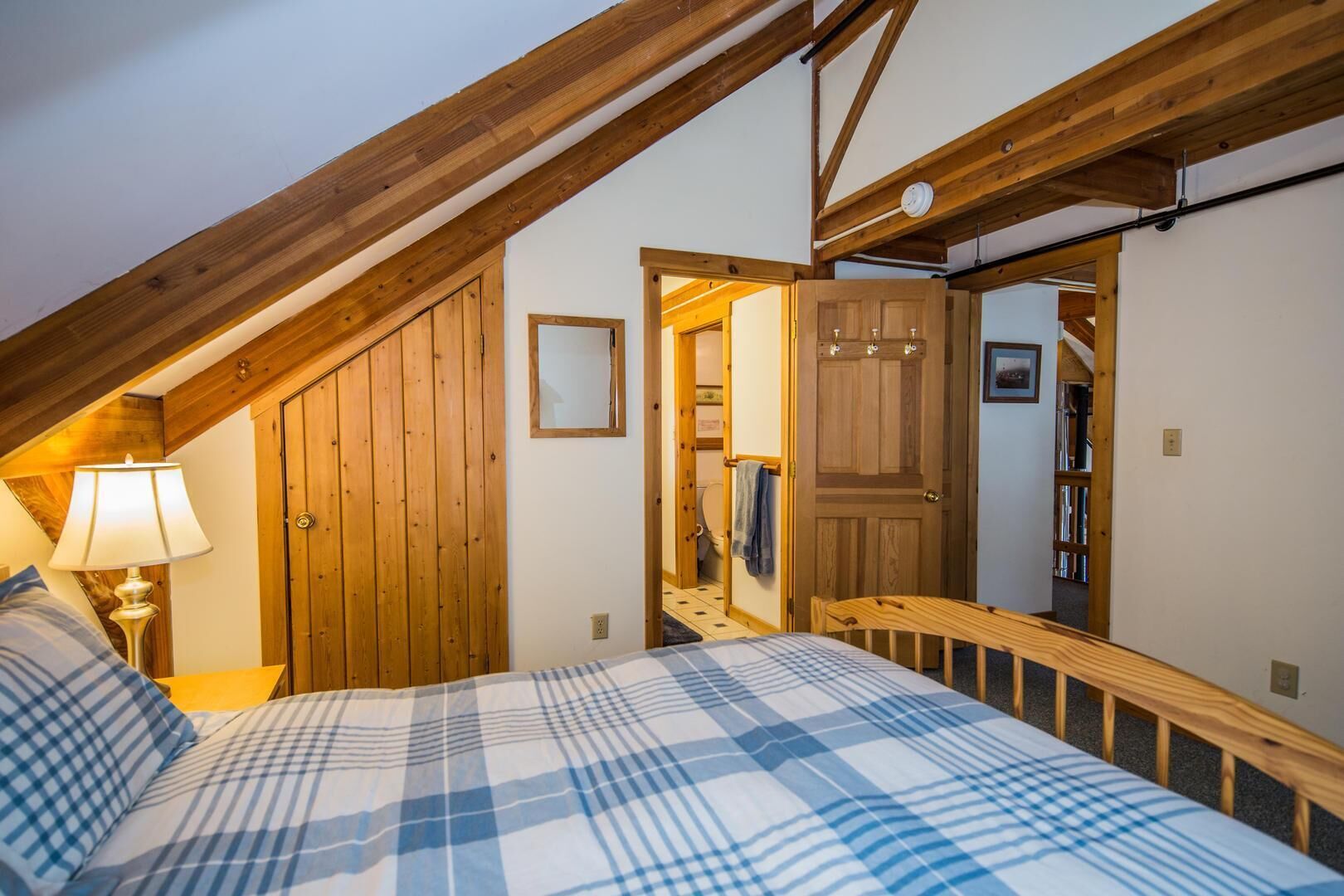 Un dormitorio con techo inclinado, vigas de madera, una cama con colcha escocesa, una mesita de noche con lámpara, una puerta de madera y un espejo en la pared.