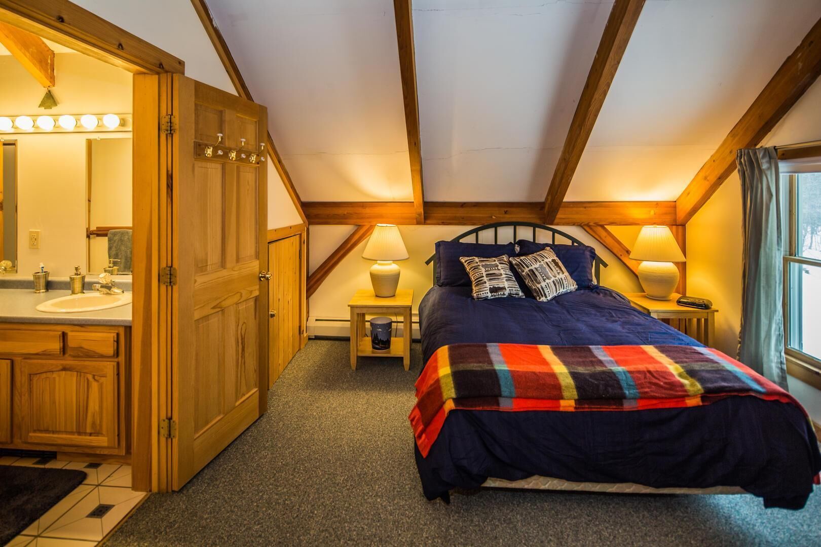 A bedroom with a bed, a nightstand, a lamp, a window, and a bathroom with a sink and mirror.