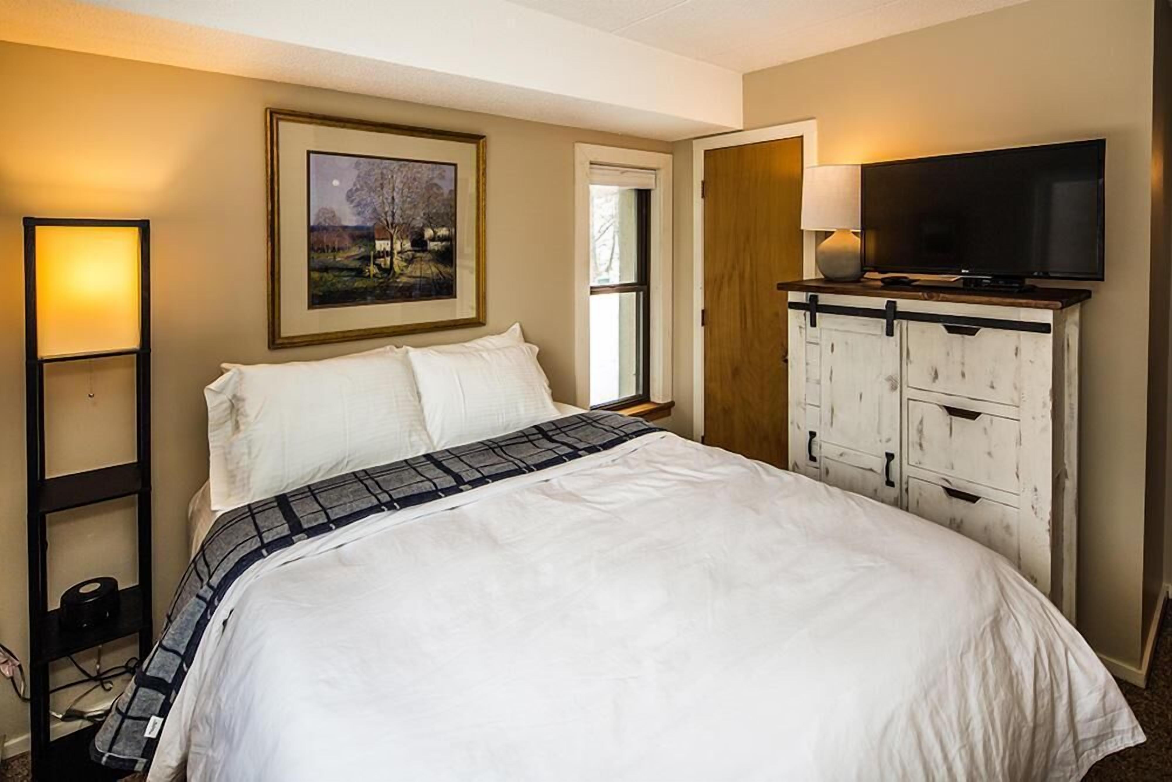 A bedroom with a bed, a TV on a dresser, a painting on the wall, and a window.