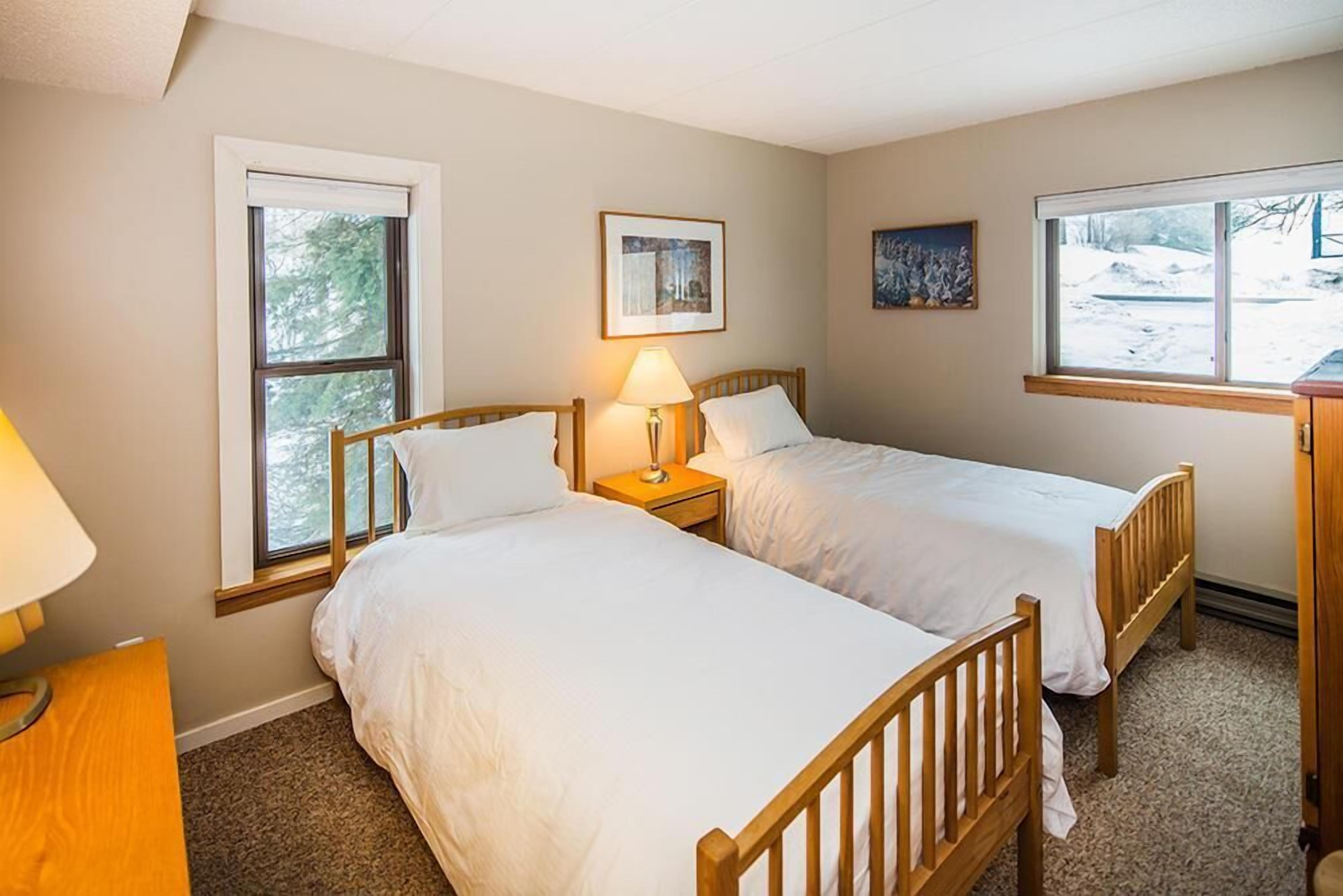 A bedroom with two single beds, a nightstand with a lamp, and a window with a view of trees.
