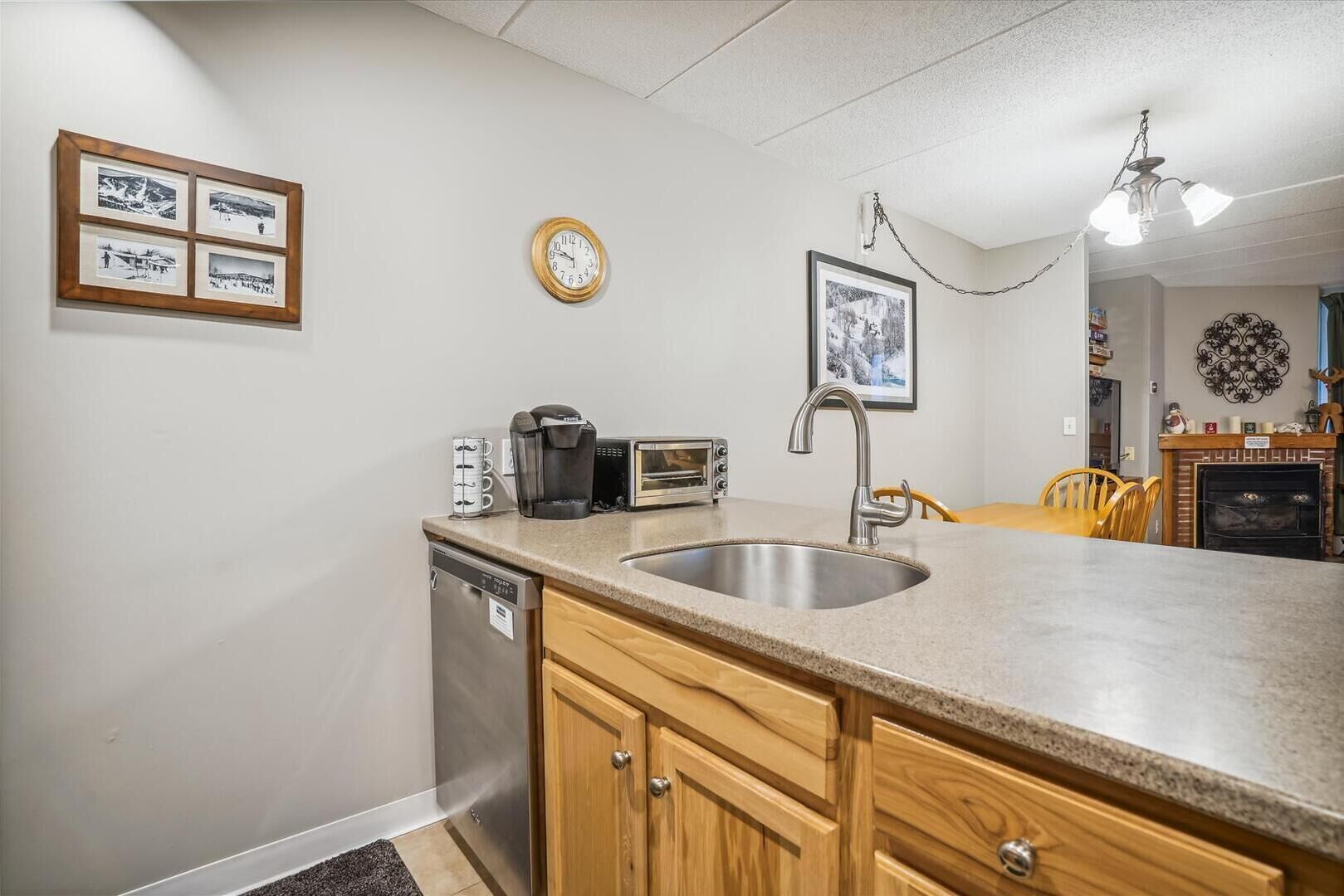 A kitchen with a dishwasher, coffee maker, toaster, and a sink. There are framed pictures on the wall and a clock.