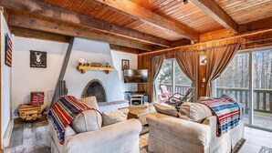 A cozy cabin interior with a fireplace, two sofas, a TV, and a large window overlooking a deck. - Hemlock Ridge 1D (Killington)