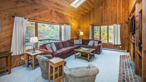 A cozy cabin interior with a wooden ceiling, large windows, a sectional sofa, a coffee table, and a fireplace. - Hideout Chalet (Killington)