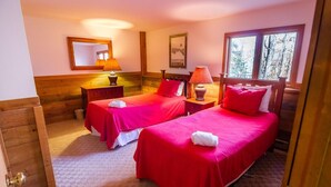 A room with two beds, each with red bedding and white towels. There are wooden headboards and nightstands with lamps. A mirror hangs on the wall, and a window provides a view of trees. - Hideout Chalet (Killington)