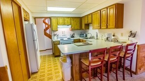 A compact kitchen with wooden cabinets, a central island with bar stools, a refrigerator, and a microwave. - Hideout Chalet (Killington)