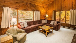 A cozy cabin interior with a wooden ceiling, a sectional sofa, a coffee table, and large windows offering views of autumn foliage. - Hideout Chalet (Killington)