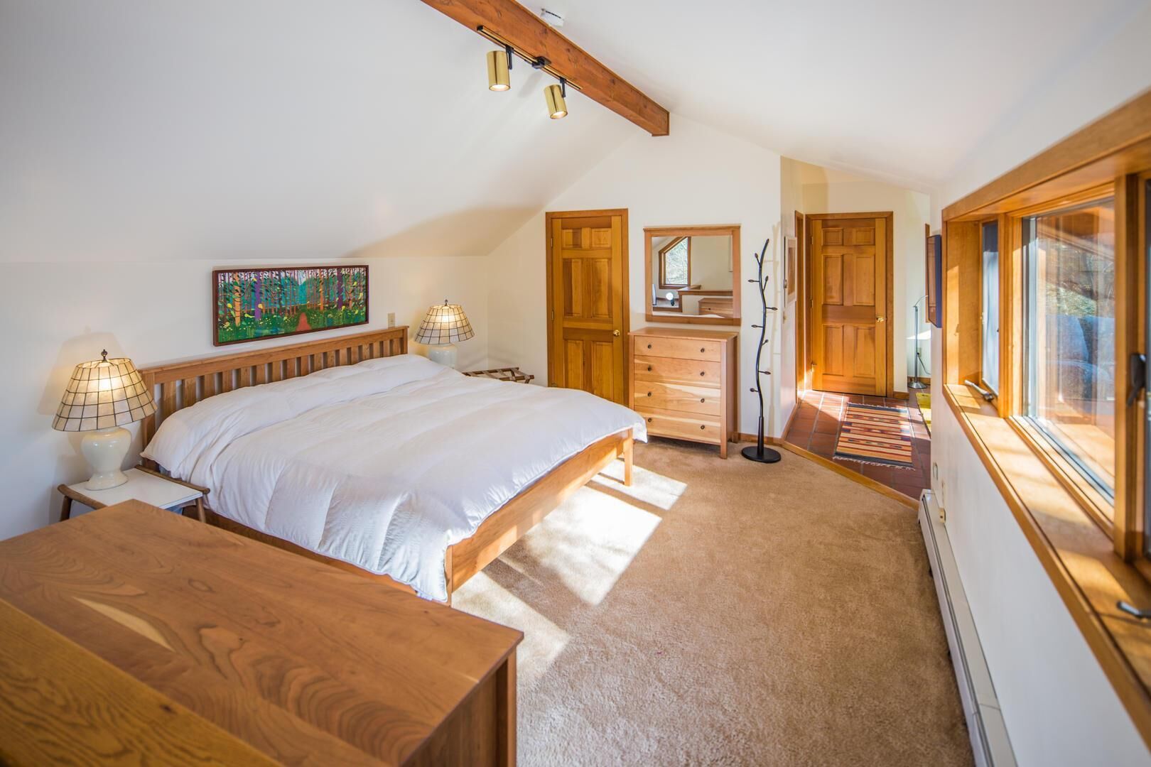 A bedroom with a large bed, wooden headboard, two bedside tables with lamps, a dresser, a coat rack, and a window with a view.