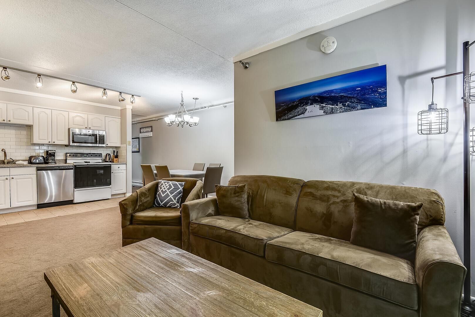 A modern living room with a brown leather sofa, a wooden coffee table, and a dining area with a table and chairs. A kitchen with white cabinets and stainless steel appliances is visible in the background.