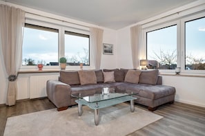 A modern living room with a glass coffee table, a brown leather sofa, and large windows with curtains.