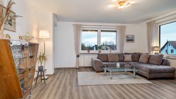 Ein modernes Wohnzimmer mit einem Ecksofa, einem Glastisch, einem Holzschrank, einer Stehlampe und einem großen Fenster mit Vorhängen.
