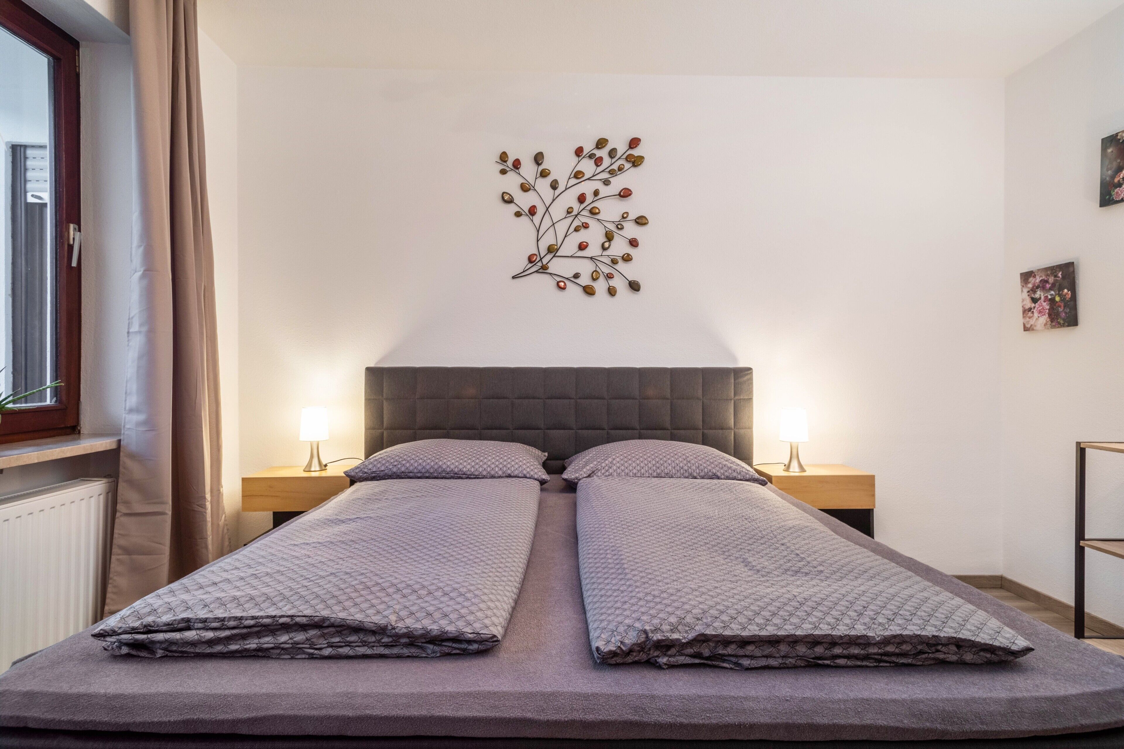 A double bed with a quilted bedspread, two bedside tables with lamps, a wall-mounted artwork, and a window with curtains.