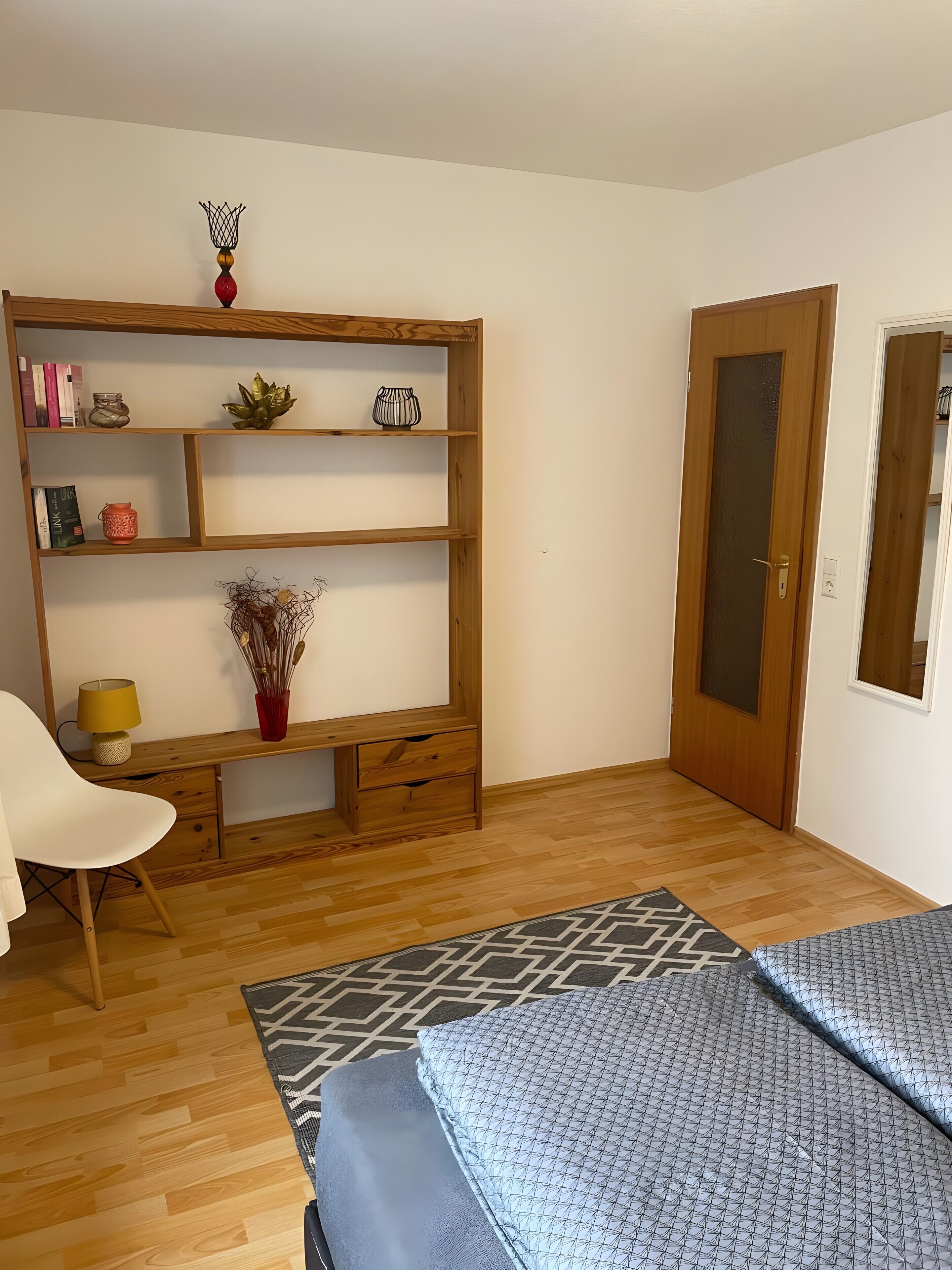 A room with a wooden bookshelf, a desk, a chair, a bed with patterned bedding, and a door.