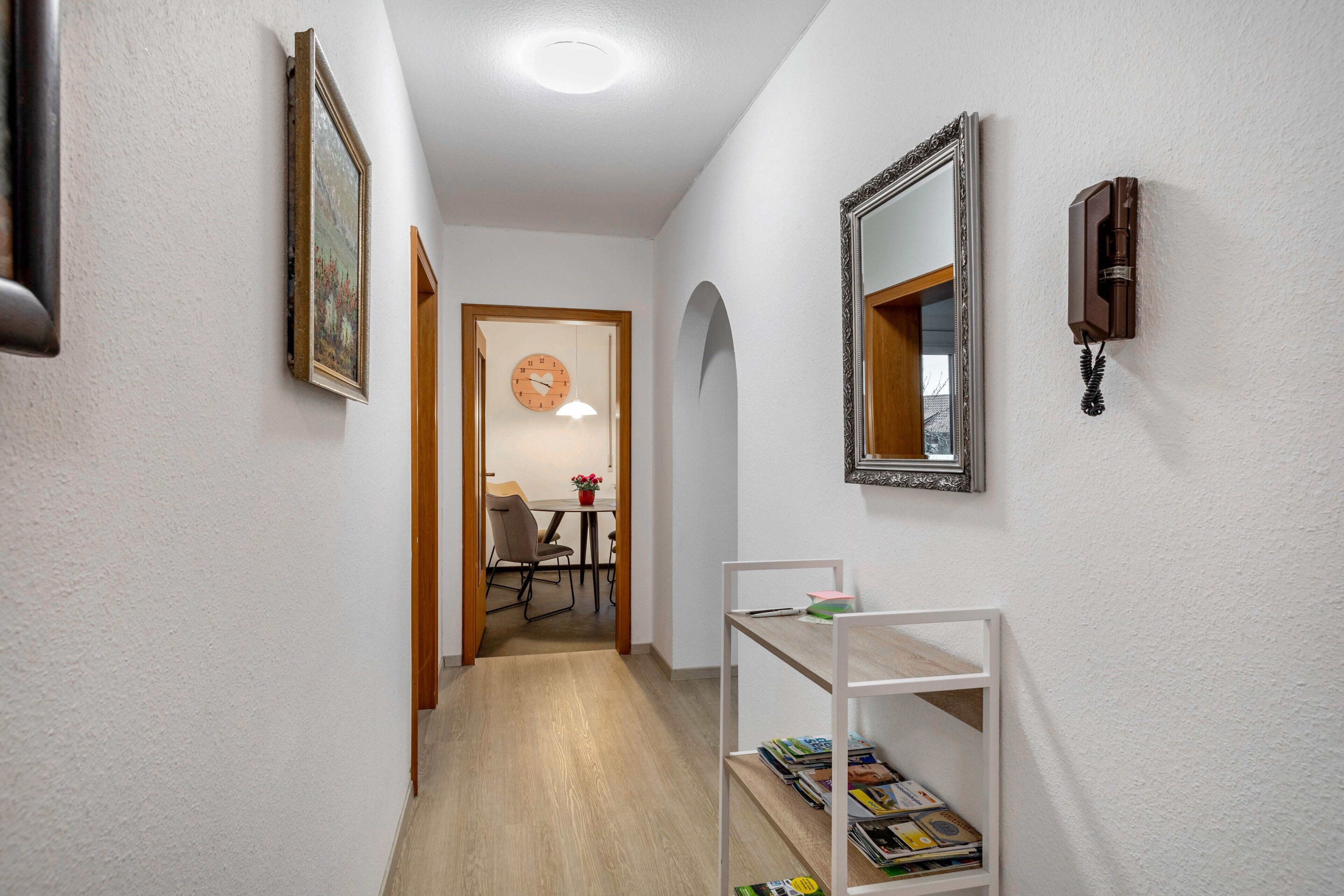A hallway with a white wall, a wooden door, a white shelf with magazines, a framed mirror, and a clock.