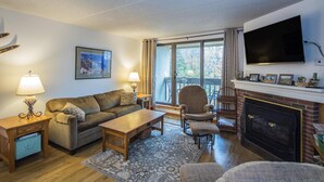 Ein gemütliches Wohnzimmer mit Sofa, Sessel, Couchtisch und Kamin. Ein Fernseher ist an der Wand montiert und es gibt ein Fenster mit Vorhängen.