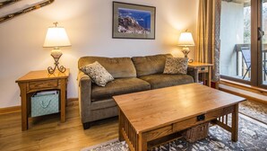 Ein gemütliches Wohnzimmer mit einer Couch, zwei Beistelltischen mit Lampen, einem Holztisch und einem großen Fenster mit Vorhängen.