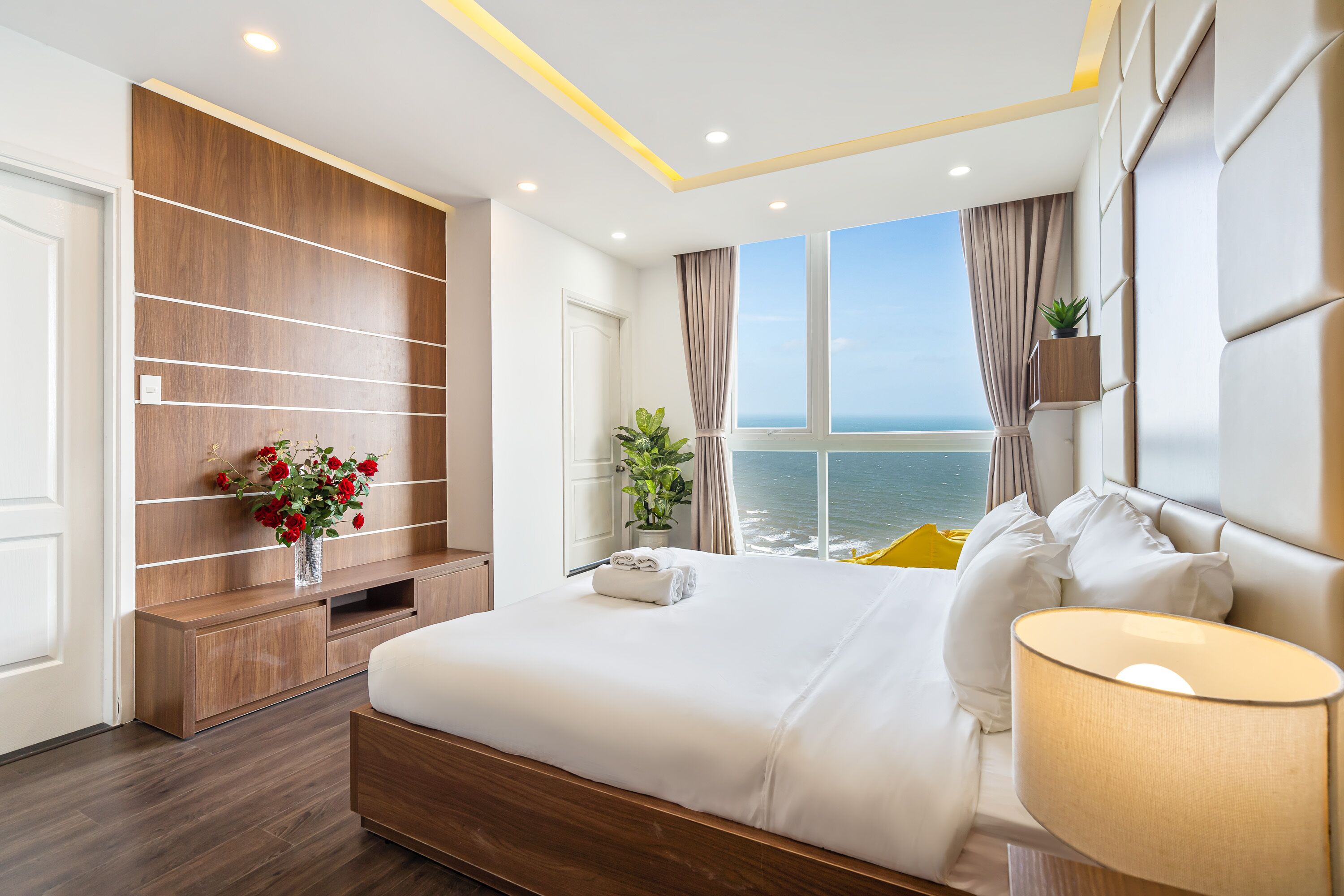 A modern hotel room with a large bed, a wooden built-in shelf with a vase of flowers, a potted plant, and a view of the sea through large windows.