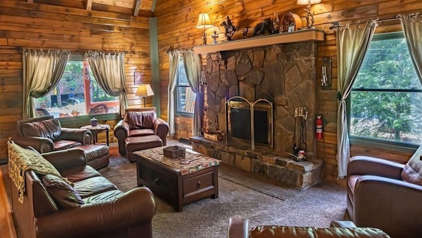 A cozy log cabin interior with leather sofas, a stone fireplace, and a large window offering a view of the outdoors.