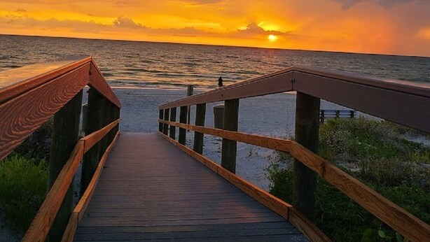 Un paseo marítimo de madera que lleva al océano, con un atardecer de fondo.