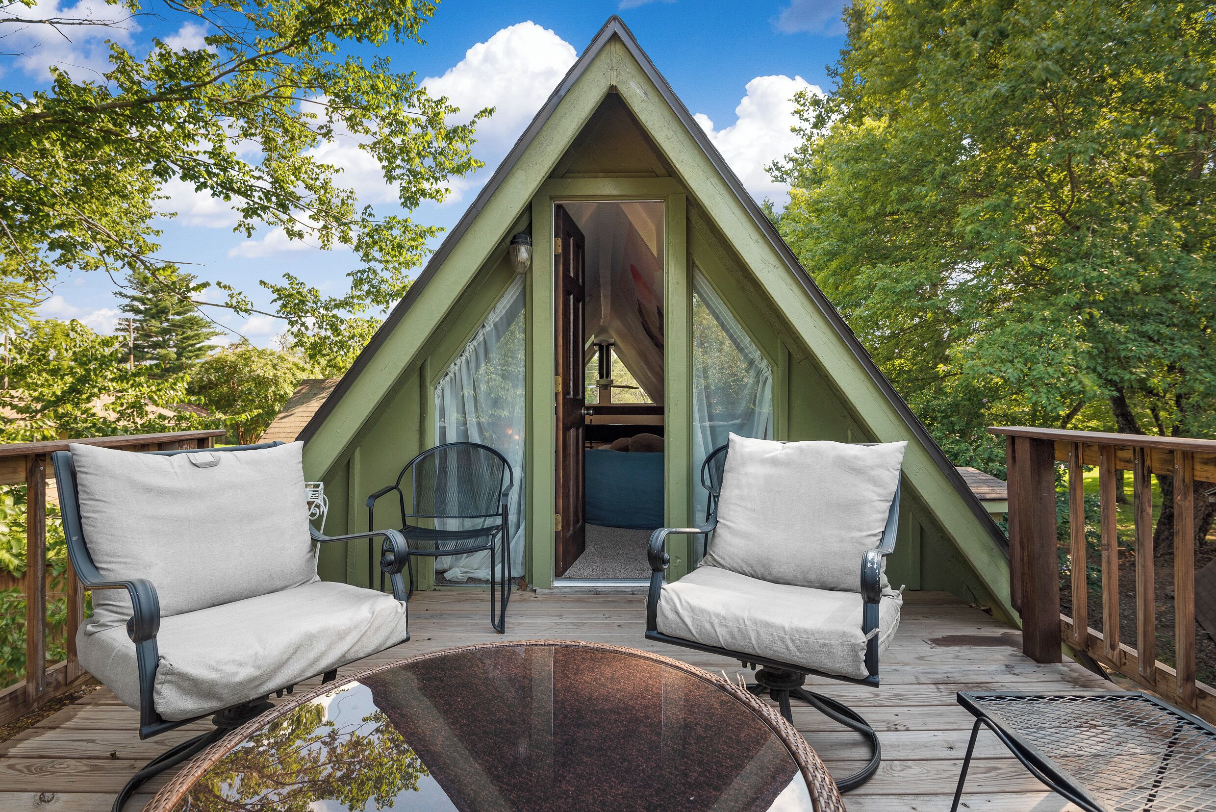 Une structure triangulaire en forme de toit à deux versants, avec deux chaises et une table sur une terrasse en bois, le tout entouré d’arbres.