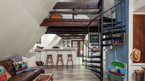 A loft-style living space with a wooden staircase, a loft bed, a kitchen area, and a dining table with stools.