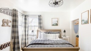 Un dormitorio con cama, mesita de noche, lámpara, ventana con cortinas, un perchero en la pared y una lámpara en el techo.