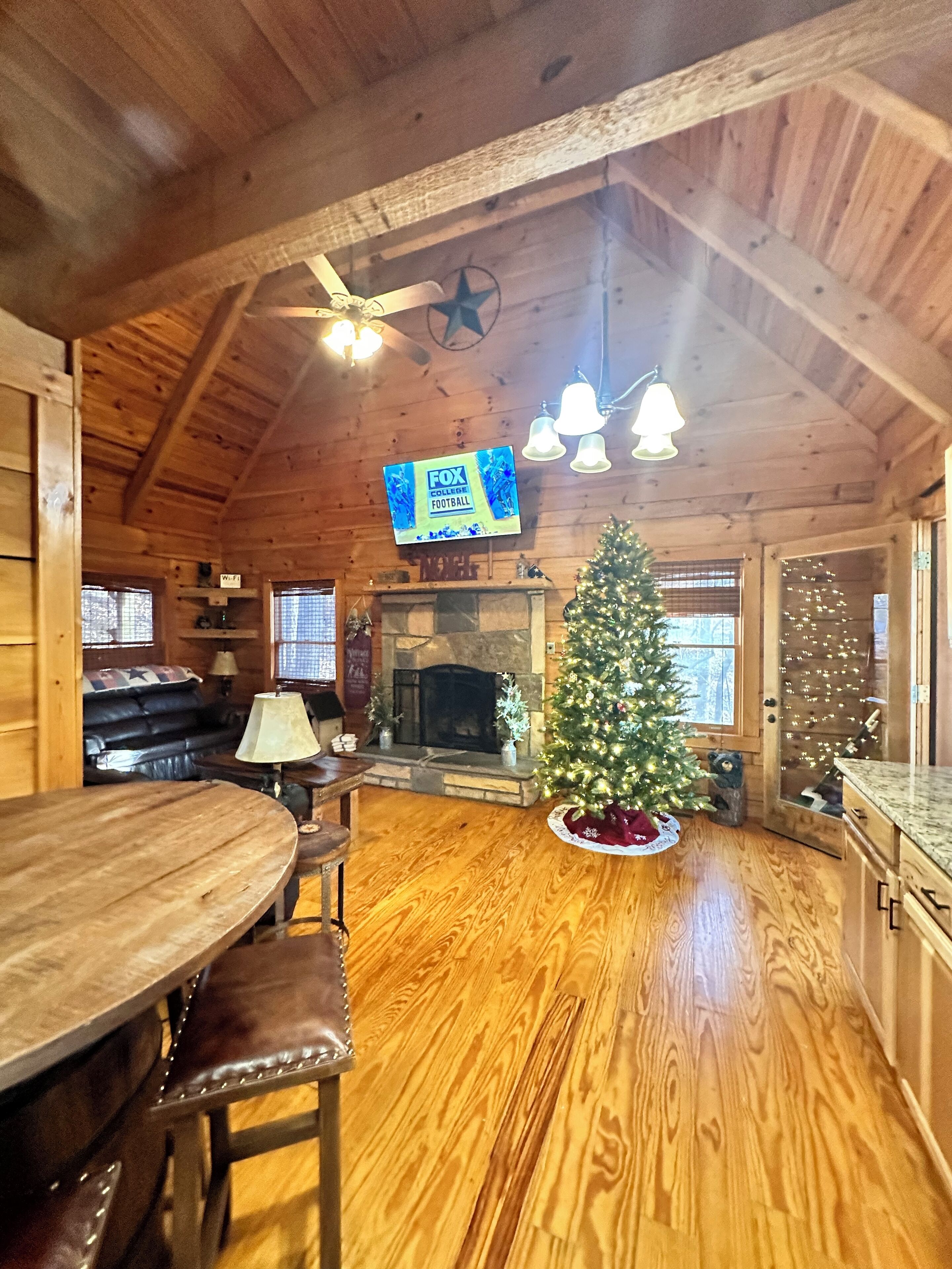 A cozy cabin interior with a fireplace, a Christmas tree, a TV mounted on the wall, and a wooden dining table.