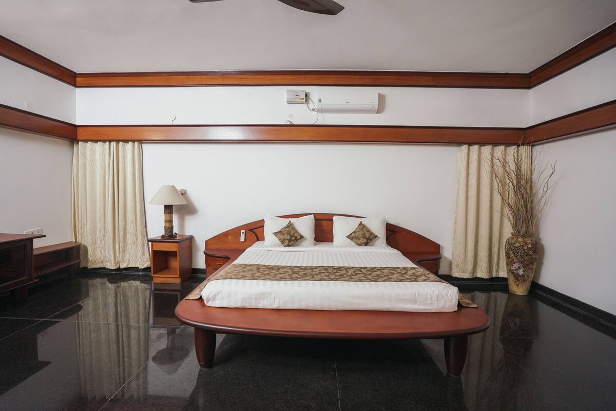 Habitación de hotel con cama de madera, mesita de noche con lámpara, un florero con plantas secas y un ventilador de techo.