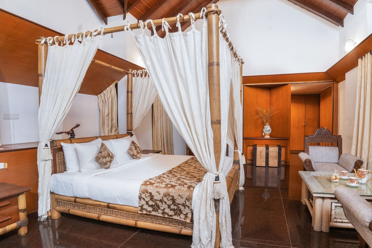 Una cama con dosel de cuatro postes con una colcha blanca y un chal floral, una mesita de noche de madera, una silla y una mesa con juego de té.