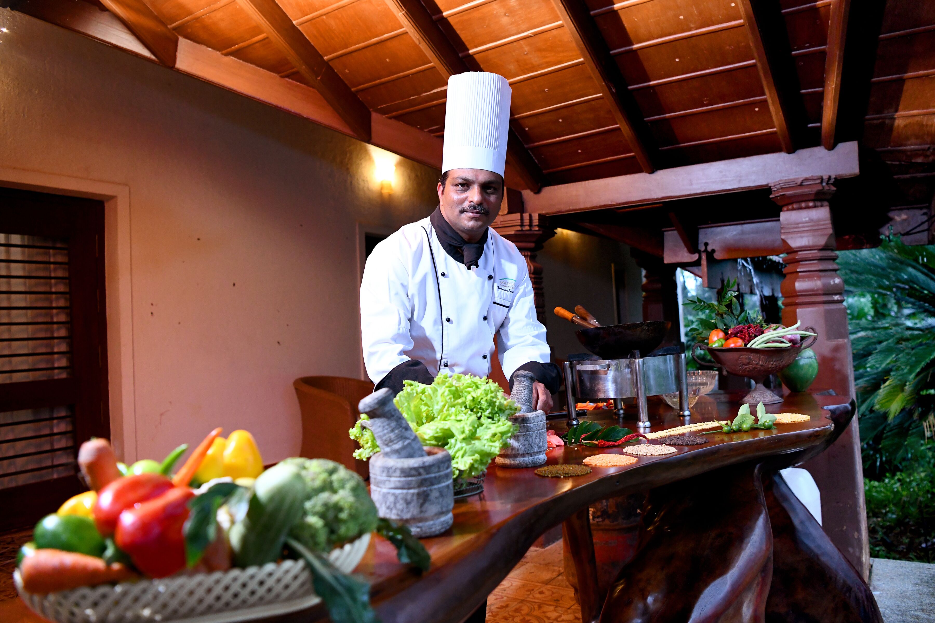 A chef in a white uniform with a tall hat, standing in a kitchen with wooden beams on the ceiling. There are various vegetables and kitchen tools on the counter.