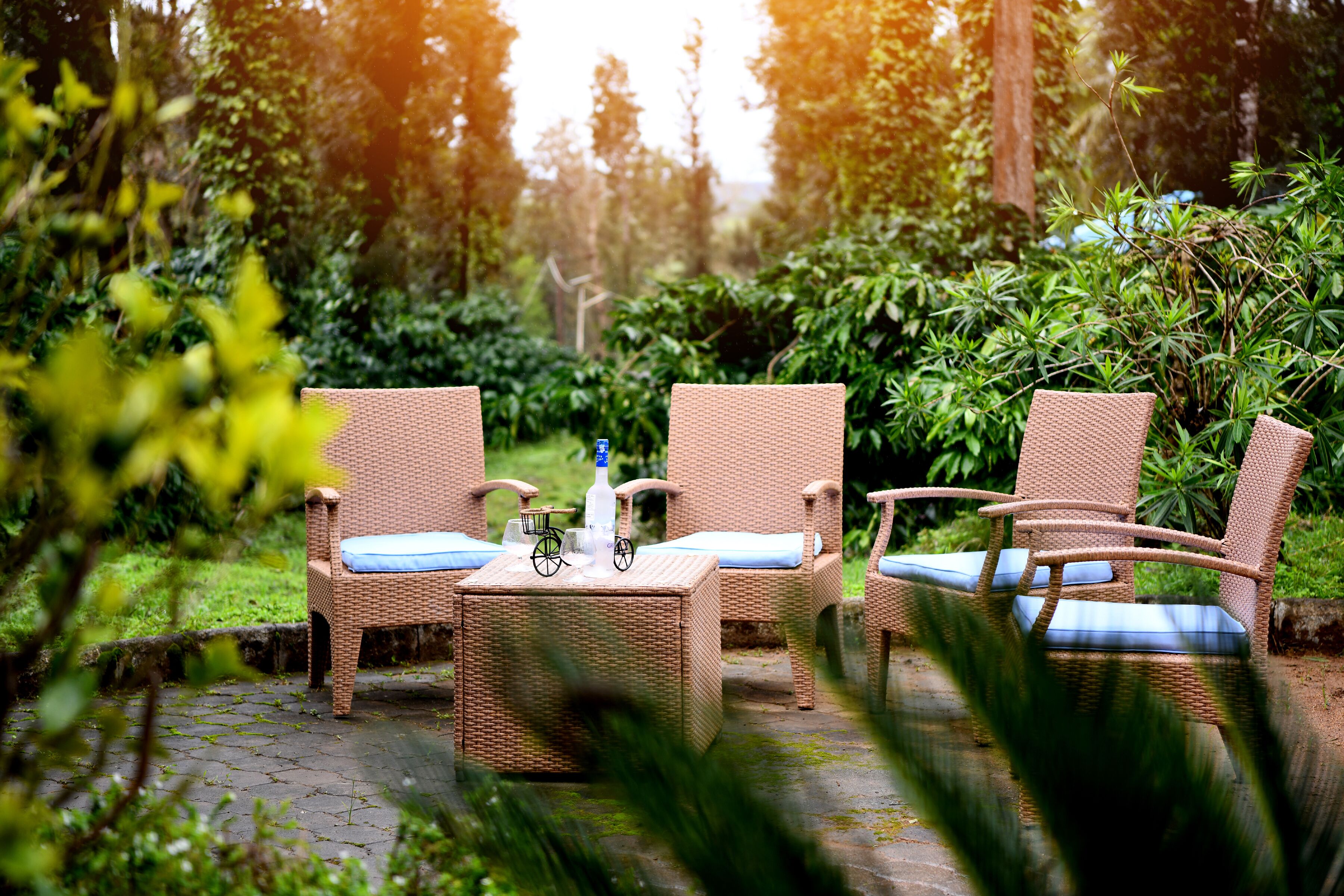 Wicker furniture with cushions, a bottle, and a bicycle wheel on a table in a garden setting.