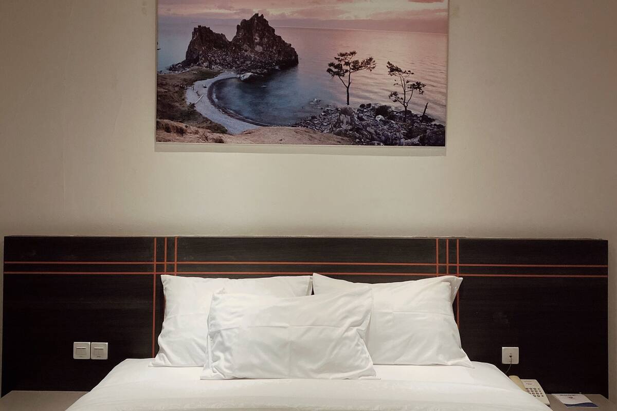 A hotel room with a large bed, a headboard with a geometric design, and a painting of a coastal landscape on the wall.